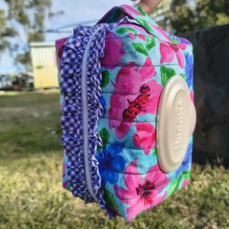 Quilted boxy nappy pouch made from blue, pink, purple and green floral fabric with a purple gingham ruffle feature along either side of the zipper.